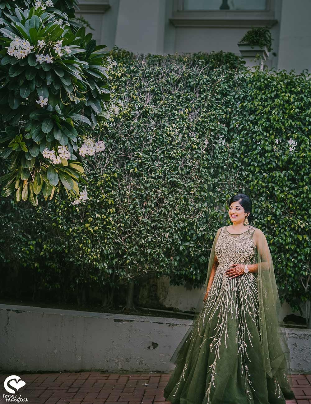 Bride Sonali in traditional attire posing against a lush green floral backdrop at her wedding