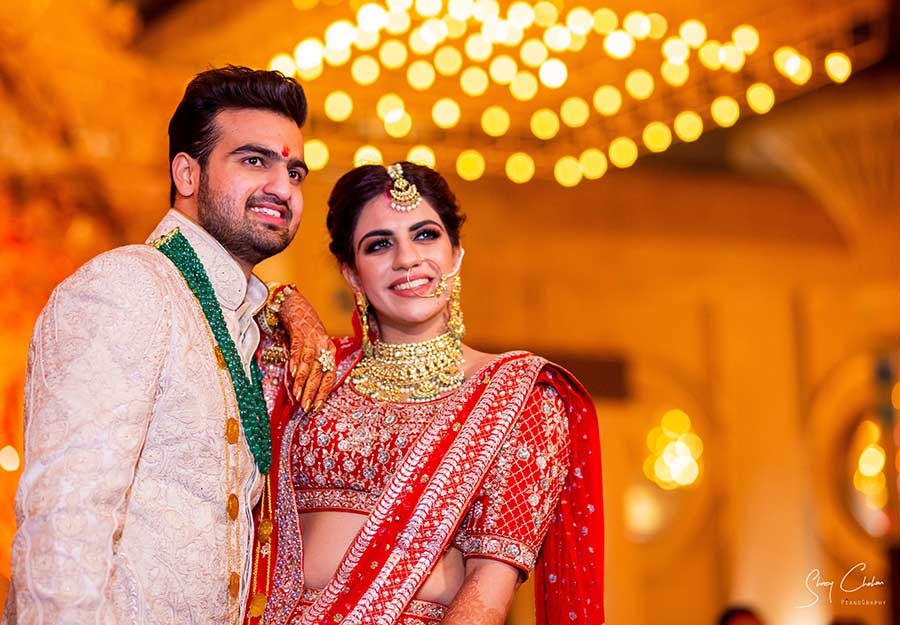 Bride and groom standing together wearing garlands with festive lights behind them